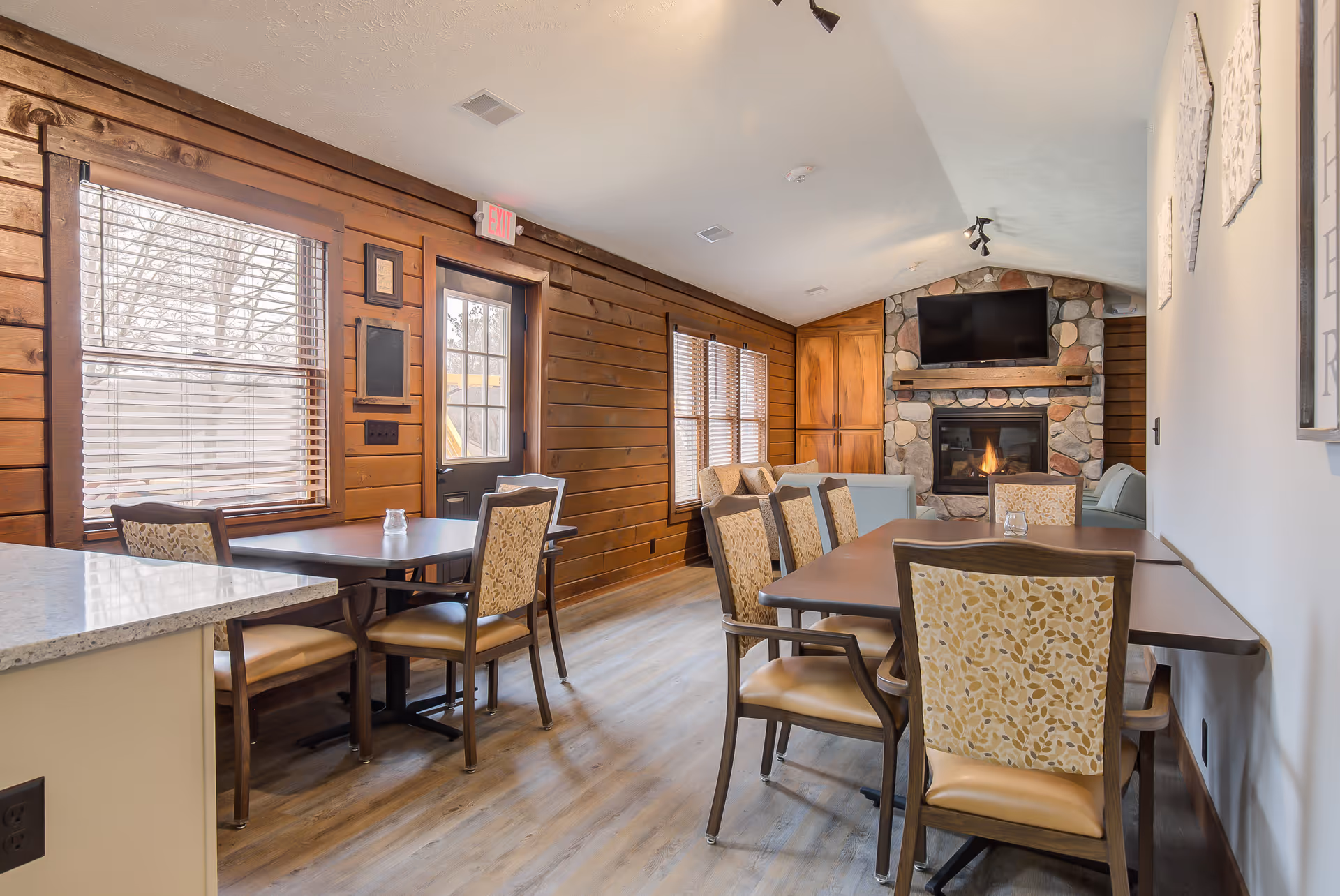 A cozy interior room with wooden walls and large windows with blinds. The room features two tables with cushioned chairs, a stone fireplace with a mounted TV above it, and light-colored wood flooring. The space is well-lit with natural light coming through the windows.