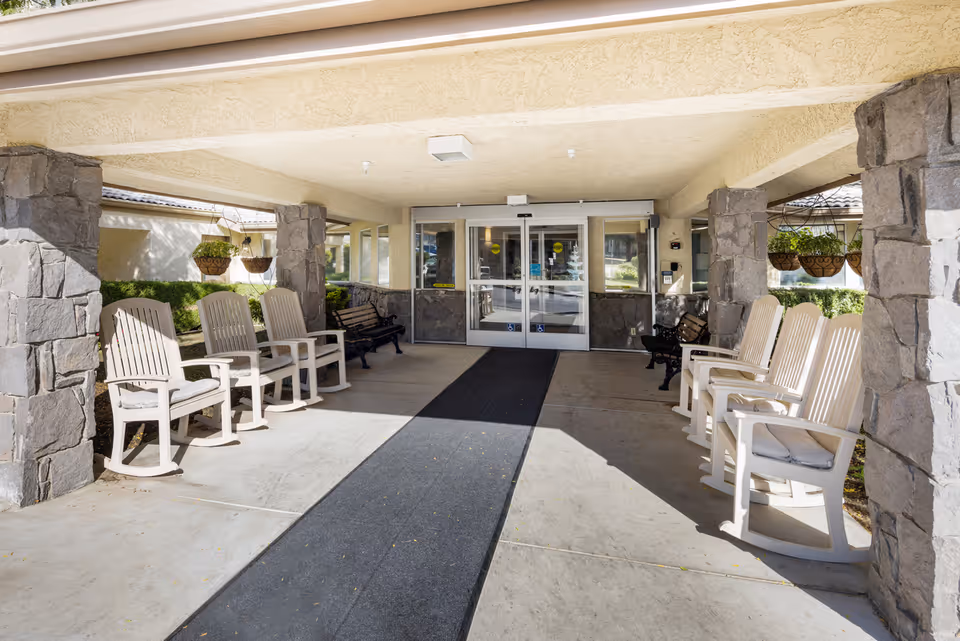 Covered entrance area of a senior living community with stone pillars and a row of white rocking chairs on each side. There are hanging flower baskets and benches near the glass double doors leading inside.