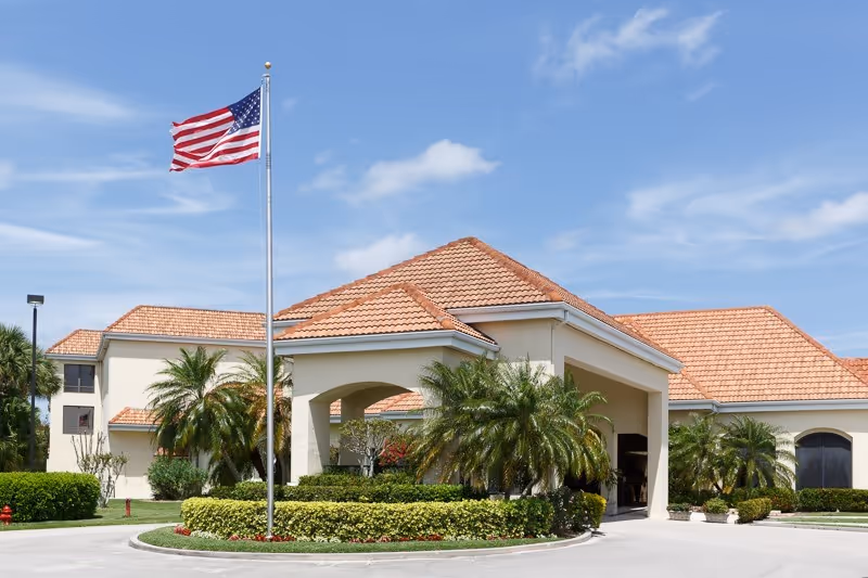 Exterior view of a senior living facility with a tiled roof, palm trees, and an American flag on a flagpole in front under a blue sky with some clouds.