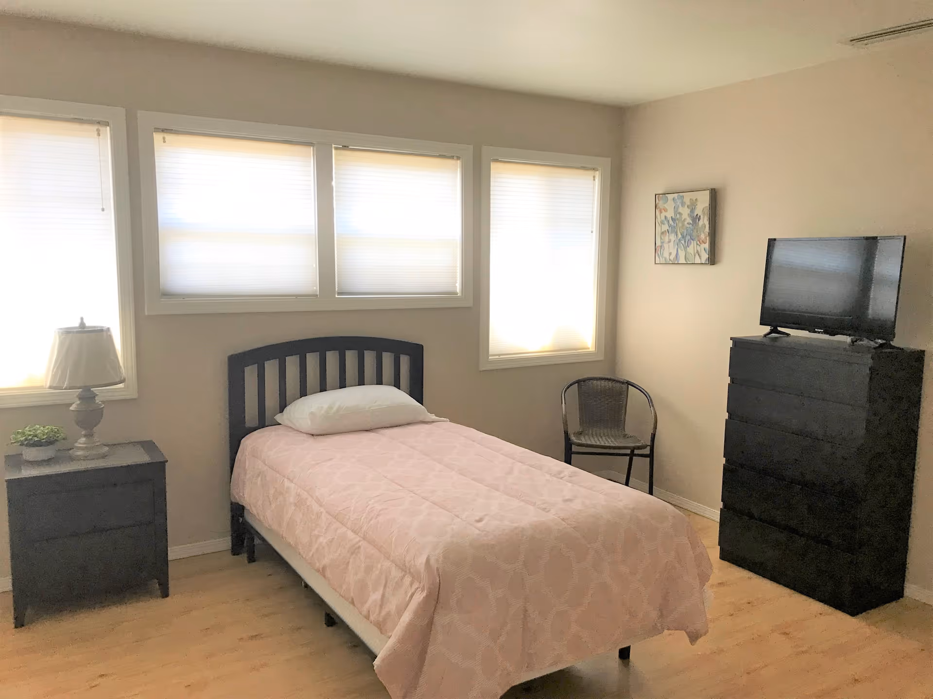 Bedroom with a single bed covered in a pink quilt, a nightstand and lamp, a black dresser with a TV, a chair, and three windows.