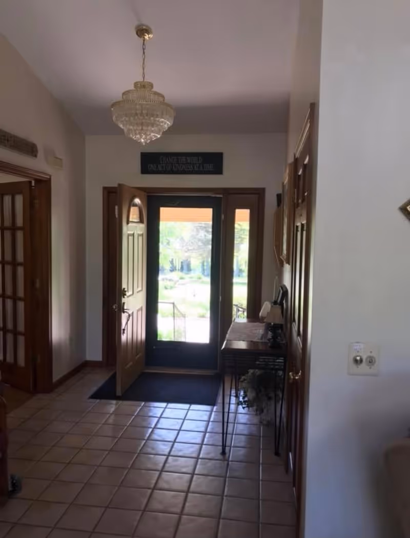 Tiled entry foyer with an open wooden front door and glass storm door, a chandelier overhead and a narrow console table along the right wall.