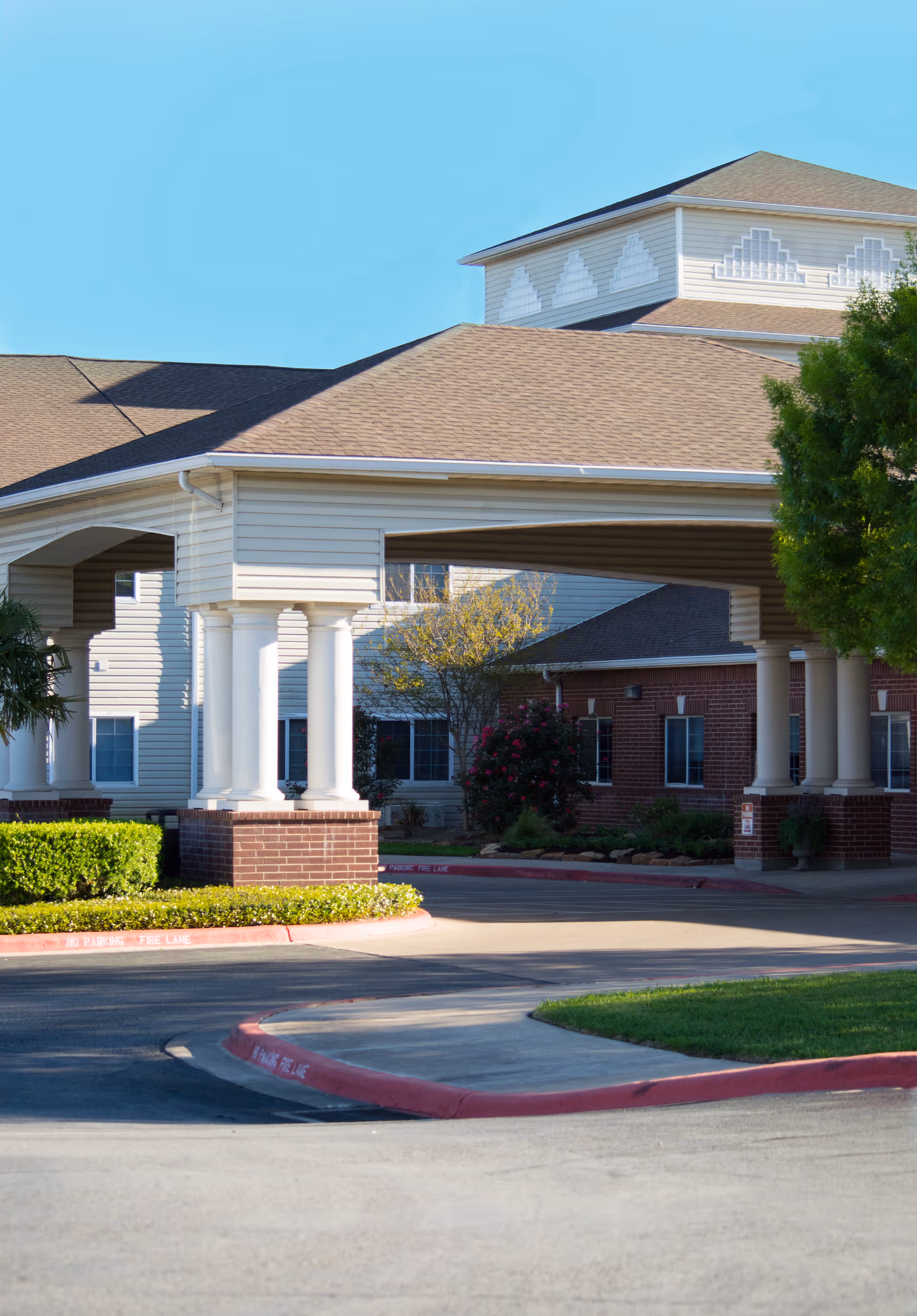 Covered porte-cochère entrance of a multi-story senior living building with white columns, brick accents, driveway, and landscaping.