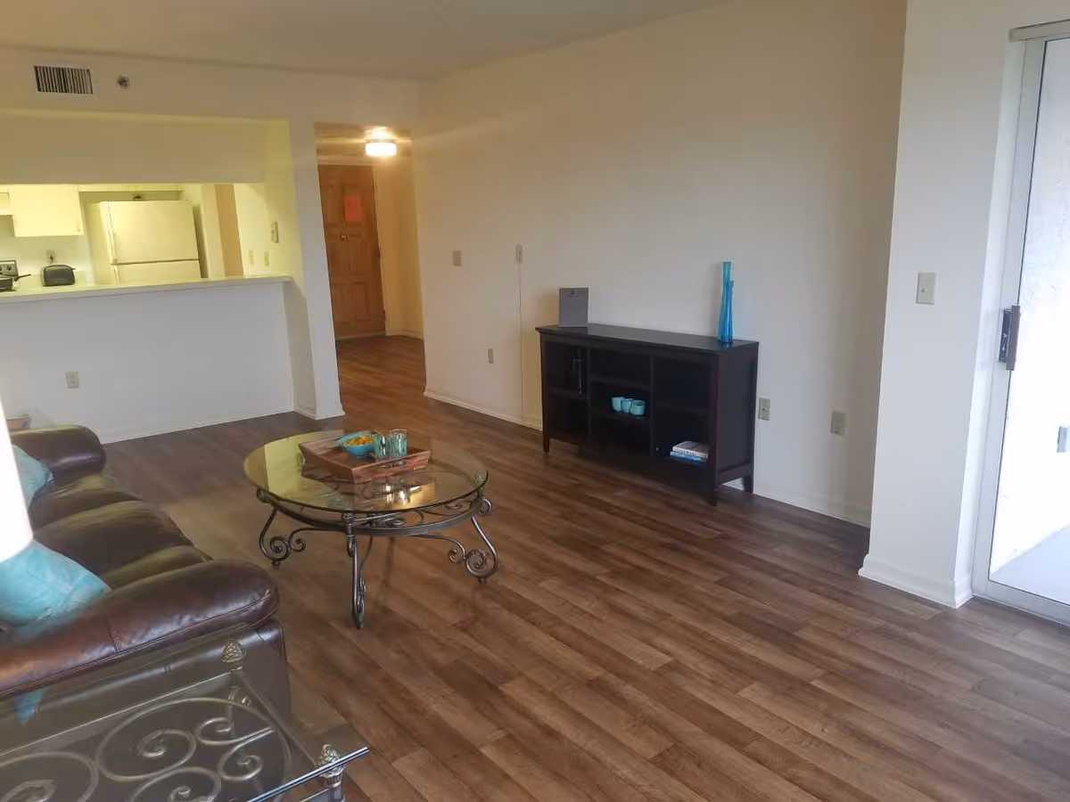 Interior view of a living room area with wooden flooring, a brown leather sofa with teal cushions, a glass coffee table with a wooden tray and cups, a dark wooden cabinet with decorative items, and a kitchen area visible in the background with white appliances. There is a door leading outside on the right side.