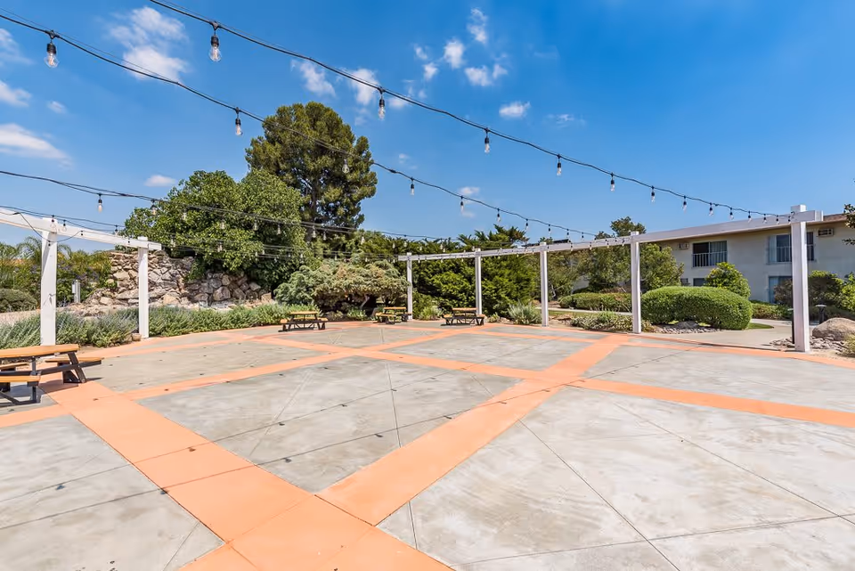 Outdoor patio area with string lights hanging overhead, picnic tables, and surrounding greenery including trees and bushes under a blue sky with some clouds.