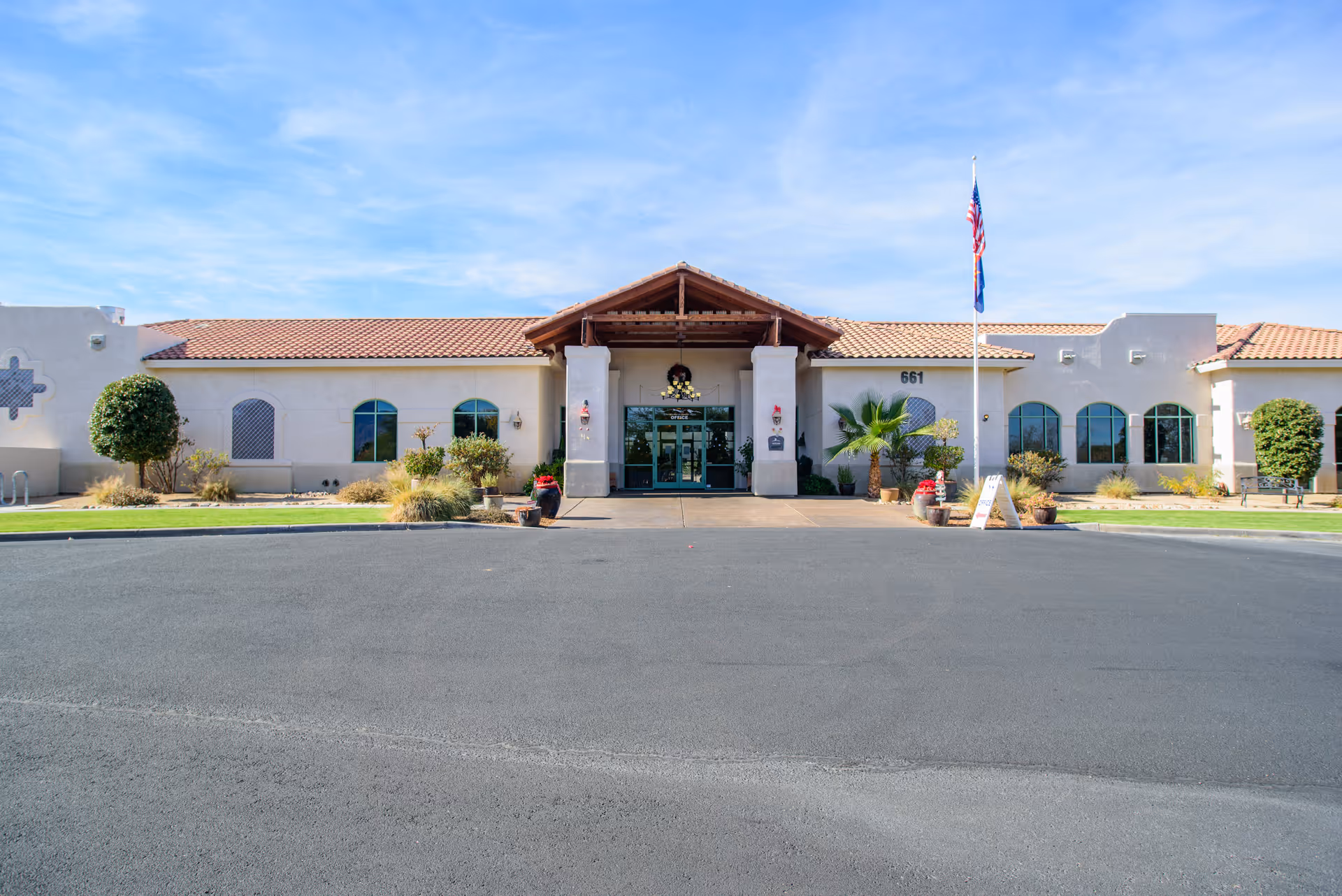Front exterior view of The Peaks At Santa Rita facility with a clear blue sky, a paved driveway, landscaped bushes and trees, an American flag on a flagpole, and a covered entrance with glass doors.