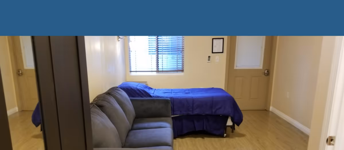 A small room with a single bed covered in a blue blanket positioned against the wall under a window with blinds. Next to the bed is a gray sofa. The room has light-colored walls and wooden flooring. There is a door with a window on the right side of the bed and a thermostat or control panel on the wall.
