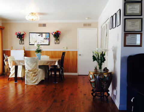 A dining room with a rectangular table covered with a beige tablecloth and surrounded by chairs, some with white covers. The room has wooden flooring and beige walls with wooden paneling halfway up. There are two wall-mounted flower arrangements on either side of a bulletin board. A small round table with a decorative vase and white flowers is positioned near the right wall, which also displays framed certificates. A closed white door is visible in the background.
