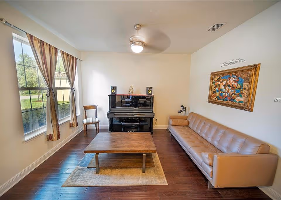 A living room with a long beige leather sofa on the right wall, a wooden coffee table on a small rug in the center, and a black upright piano against the far wall with two speakers on top. There is a wooden chair with a striped cushion next to the piano. Two large windows with striped curtains are on the left wall, letting in natural light. A colorful framed painting hangs above the sofa, and a ceiling fan with a light is on the ceiling.