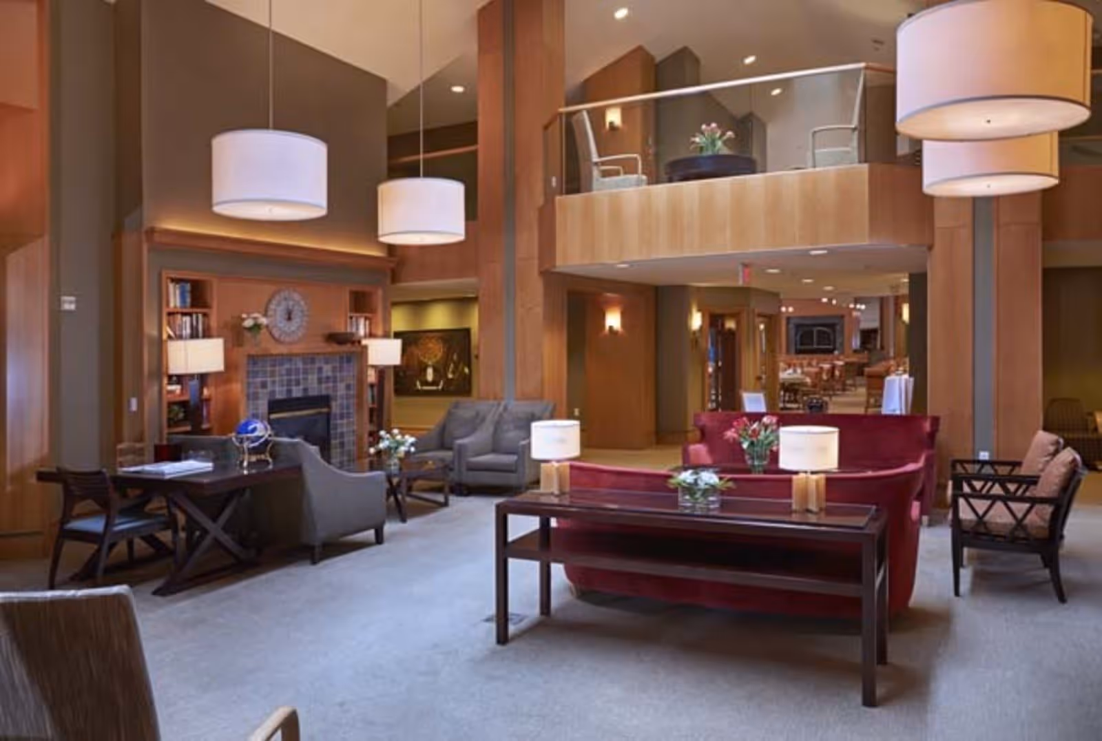 A spacious and well-lit senior living facility common area with high ceilings, modern pendant lights, comfortable seating including red and gray sofas and armchairs, a fireplace with a clock above it, and decorative flowers on tables. There is a balcony area with additional seating overlooking the main room.