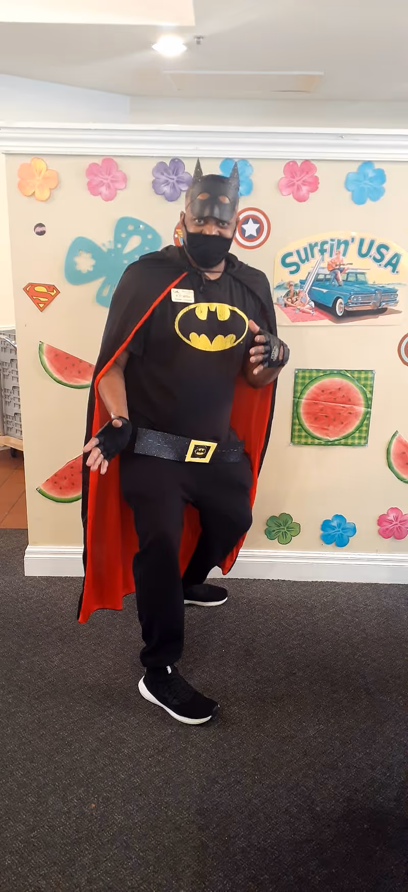 A person dressed in a Batman costume with a black mask, cape with red lining, and Batman logo on the chest, posing indoors in front of a wall decorated with colorful flowers, watermelon images, and a 'Surfin' USA' sign featuring a vintage car and beach scene.