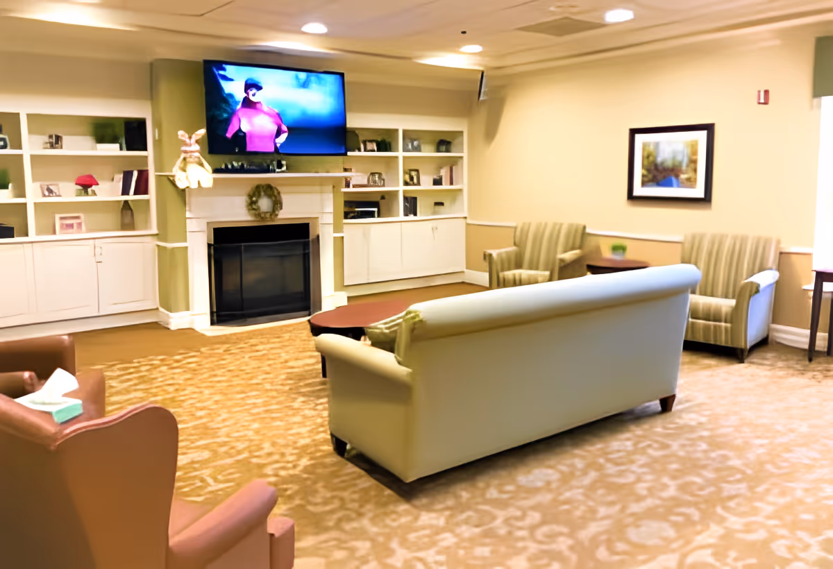 A cozy living room area in a senior living facility with a beige couch, two striped armchairs, and a brown armchair arranged around a coffee table. A fireplace with a wreath above it is centered on the far wall, flanked by built-in white shelves and cabinets. A flat-screen TV is mounted above the fireplace, and a framed picture hangs on the adjacent wall. The room has warm lighting and carpeted flooring.