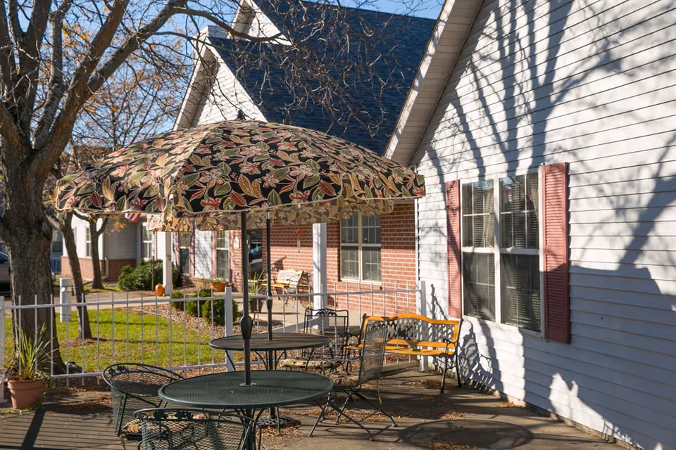 Outdoor patio with metal tables, chairs and a patterned umbrella beside the exterior of a light-colored assisted living building.