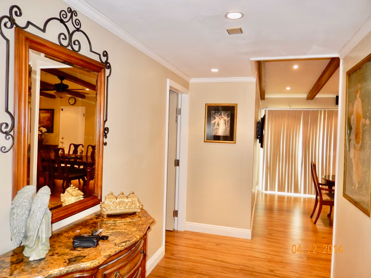 Interior hallway of a senior living facility with wooden flooring and beige walls. On the left side, there is a decorative wooden console table with a marble top, holding a small angel statue, a decorative box, and some keys. Above the table is a large mirror with an ornate frame reflecting a dining area with wooden chairs and a ceiling fan. The hallway leads to a room with vertical blinds covering a large window or door. There are framed pictures on the walls and recessed ceiling lights.