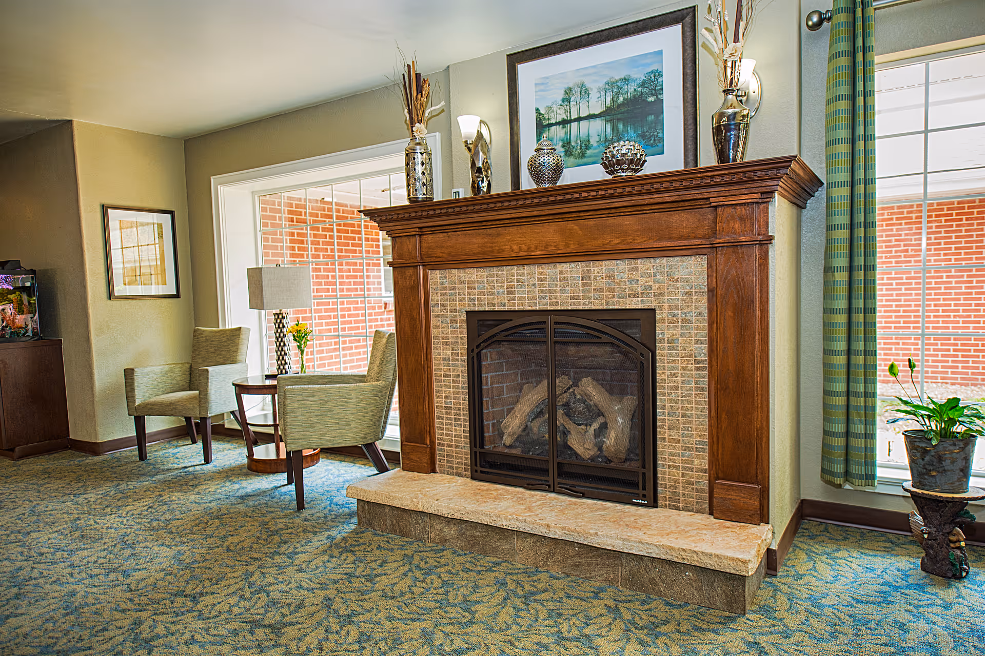 A cozy living room area featuring a wooden fireplace with a tiled surround and a stone hearth. On the mantel are decorative vases and a framed landscape painting. To the left, there are two upholstered chairs with a small round table between them, holding a lamp and a vase with yellow flowers. Large windows with green curtains allow natural light to enter the room, and a potted plant is placed near the window on the right.