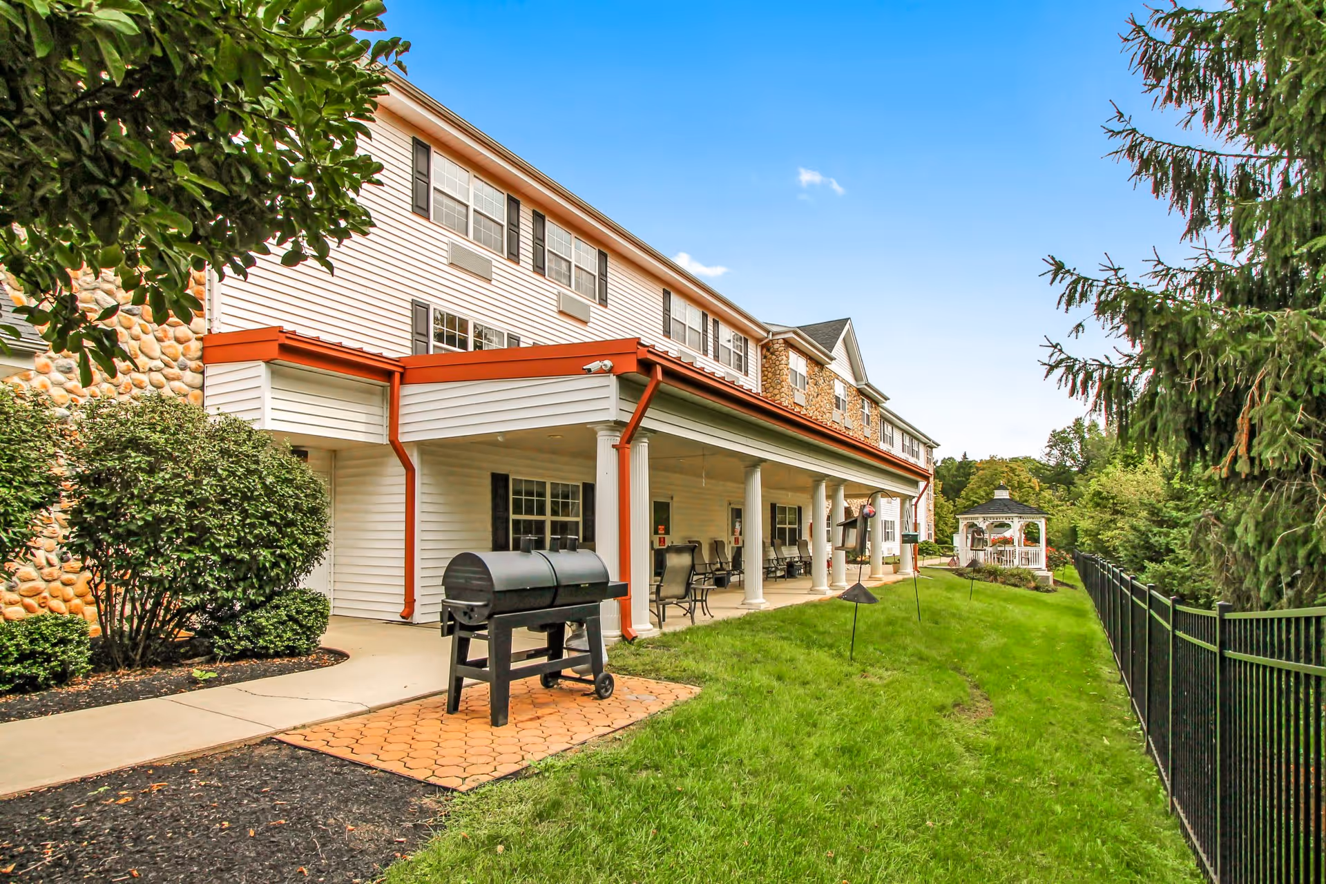 Outdoor view of a senior living facility with a covered patio area featuring chairs and tables, a black barbecue grill on a paved section, green grass, bushes, trees, and a white gazebo in the background under a clear blue sky.