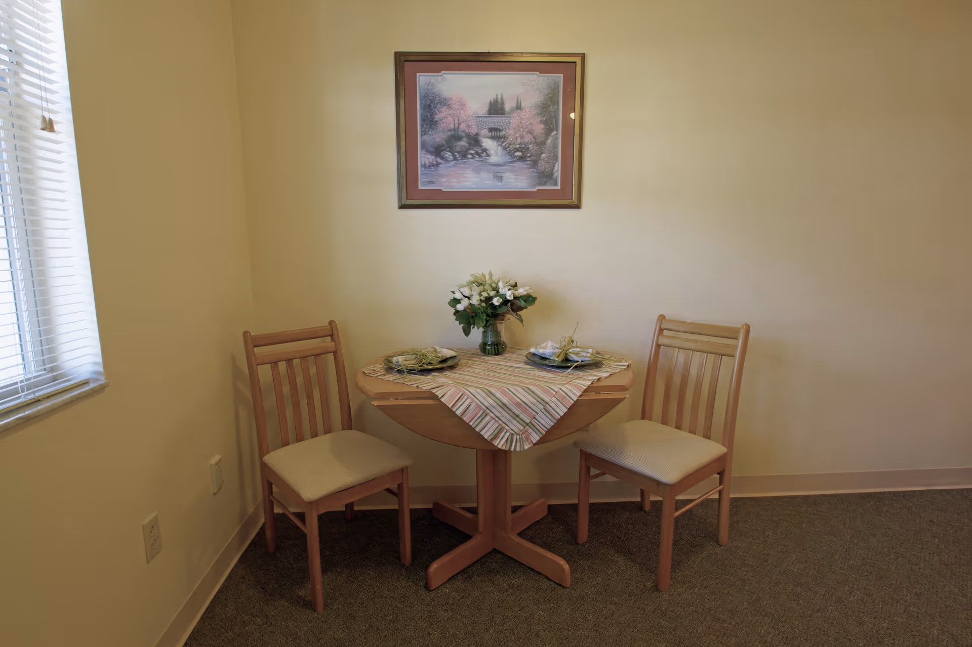 A small dining area with a round wooden table covered by a striped tablecloth and set with two place settings. Two wooden chairs with beige cushions are positioned on either side of the table. A vase with white flowers is centered on the table. A framed picture of a scenic landscape hangs on the beige wall behind the table. A window with white blinds is on the left side of the image.