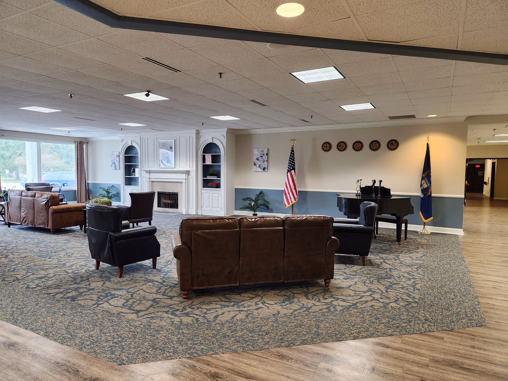 Spacious senior living common lounge with leather sofas, armchairs, a fireplace, piano, and an American flag.