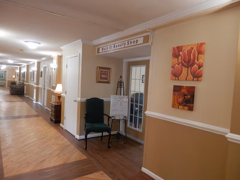 Interior hallway of a senior living facility with a 'Main St Beauty Shop' doorway, a chair, and wall artwork.