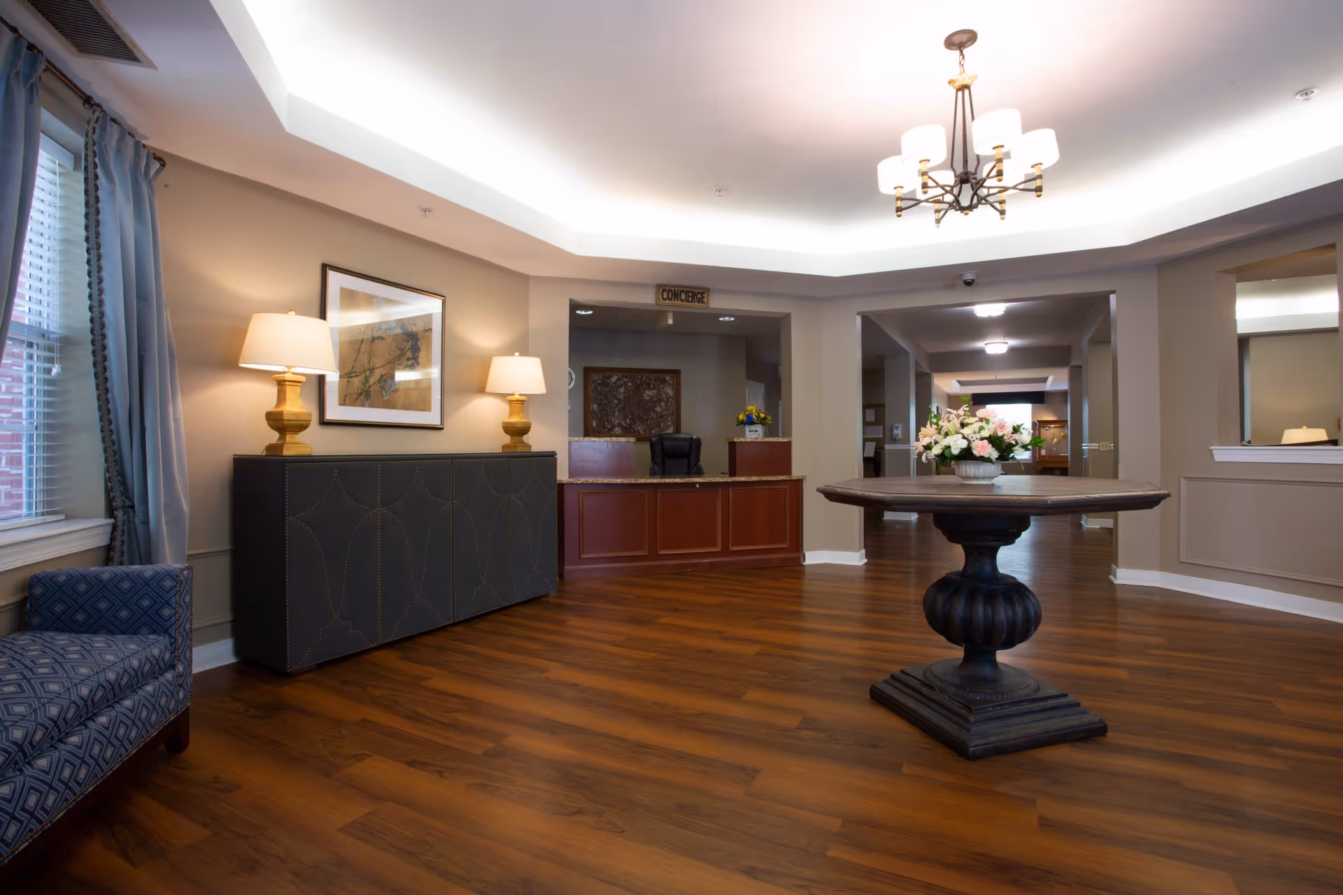 Interior view of a senior living facility lobby with wooden flooring, a round wooden table with a floral arrangement in the center, a reception desk labeled 'Concierge' in the background, two table lamps on a sideboard, a framed artwork on the wall, and a patterned armchair near a window with blue curtains.