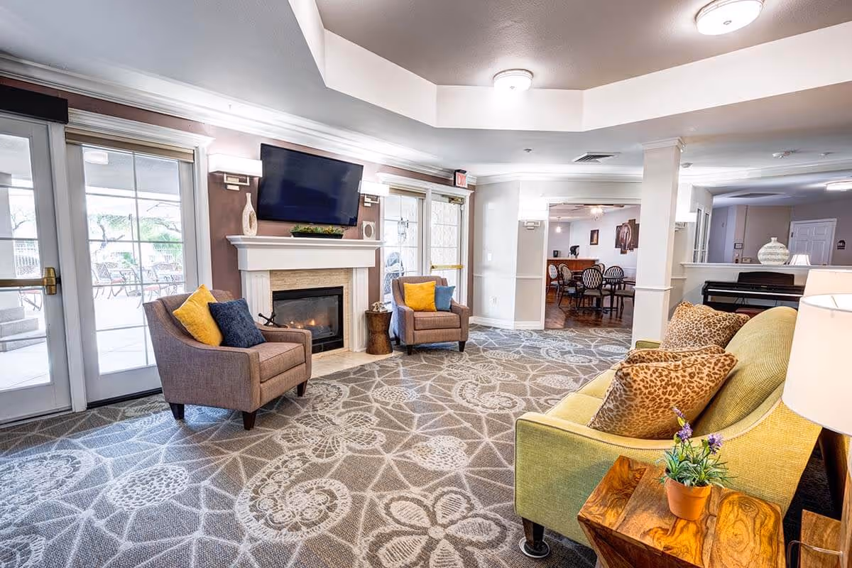 A bright and spacious living room area in an assisted living facility featuring a patterned carpet, a green sofa with leopard print pillows, two armchairs with yellow and blue cushions, a fireplace with a mounted flat-screen TV above it, and glass doors leading to an outdoor patio with seating.