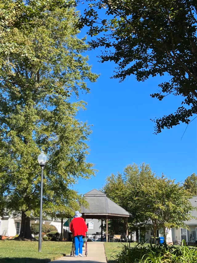A person in a red jacket using a walker walks on a sidewalk toward a gazebo in a grassy courtyard with trees under a clear blue sky.