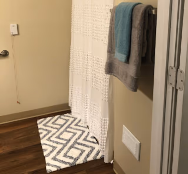A bathroom corner featuring a white textured shower curtain, a patterned blue and white bath mat on a wooden floor, and two towels hanging on a wall-mounted rack, one gray and one teal. A light switch and a vent are visible on the beige walls.