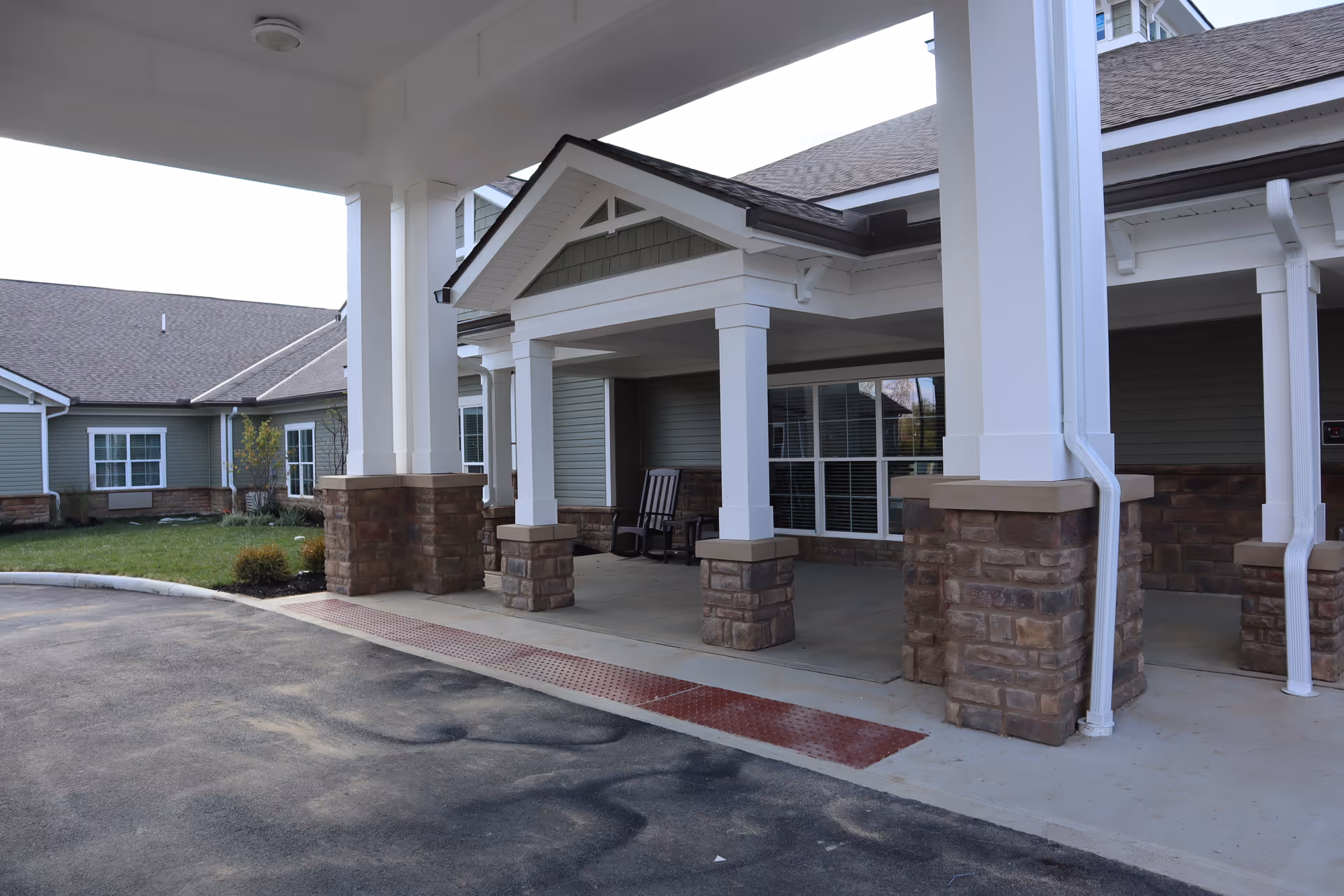 Covered entrance area of a senior living facility with white pillars on stone bases, a small porch with two rocking chairs, and green siding on the building exterior.