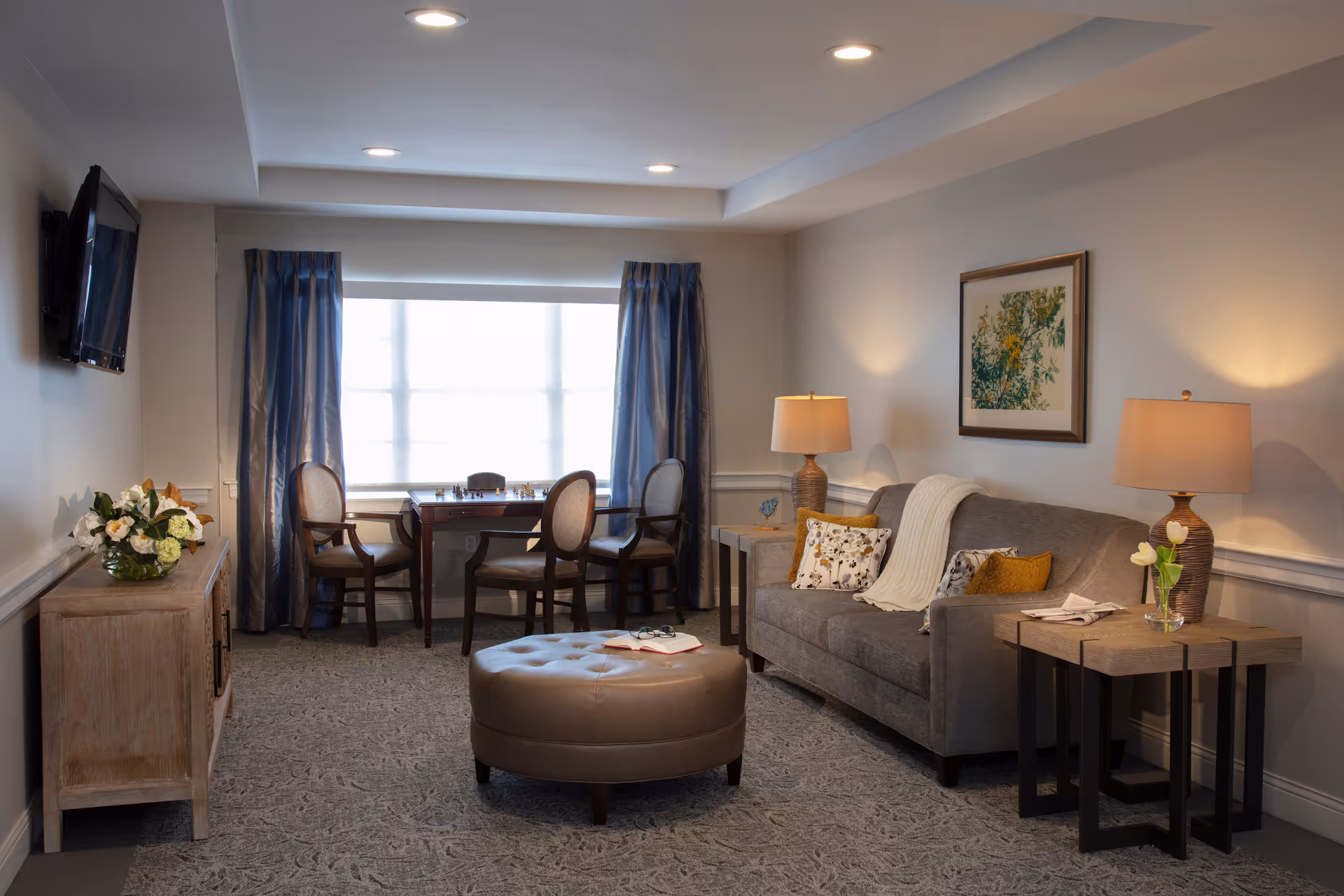A cozy living room in an assisted living facility featuring a gray sofa with decorative pillows and a throw blanket, two wooden side tables with lamps and flowers, a round leather ottoman with an open book and glasses on it, a wooden cabinet with a flower arrangement, a wall-mounted TV, and a window with blue curtains behind a small table with four chairs.