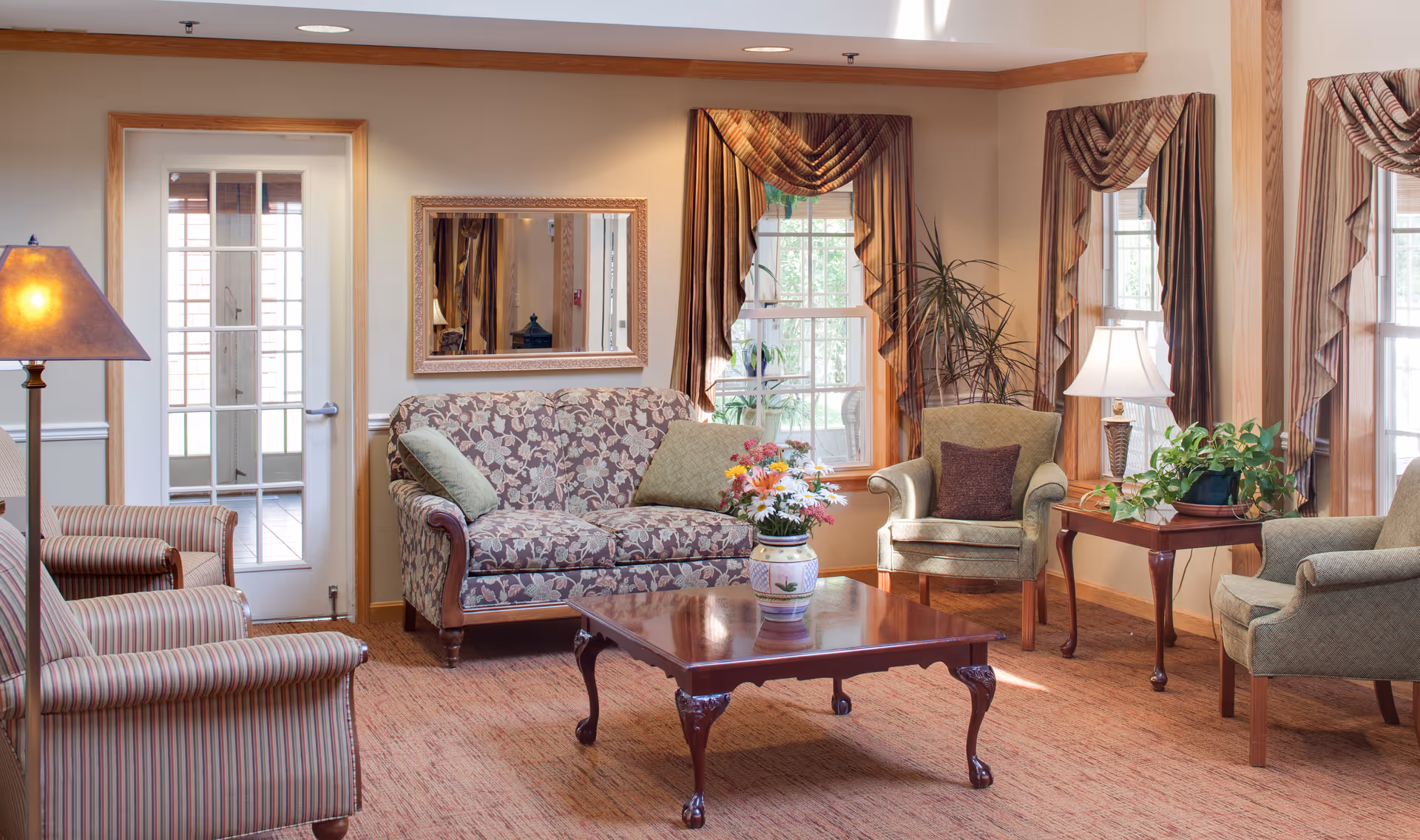 A cozy living room area in a senior living facility with a floral patterned loveseat, two green armchairs, and two striped armchairs arranged around a polished wooden coffee table with a flower vase. The room has large windows with striped curtains, a floor lamp, a table lamp on a side table with a potted plant, and a wall mirror above the loveseat.