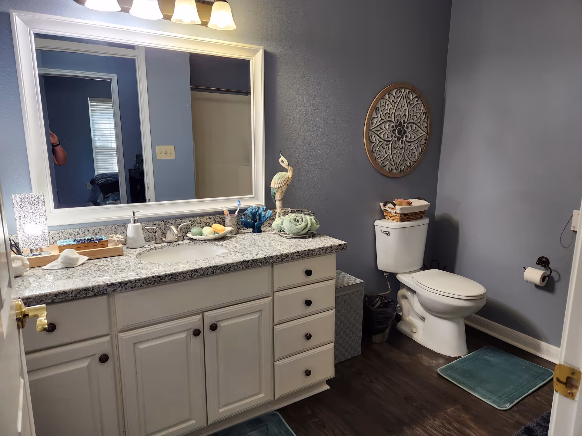 Well-lit bathroom with a white granite vanity and large mirror, decorative wall art, and a toilet against blue-gray walls.