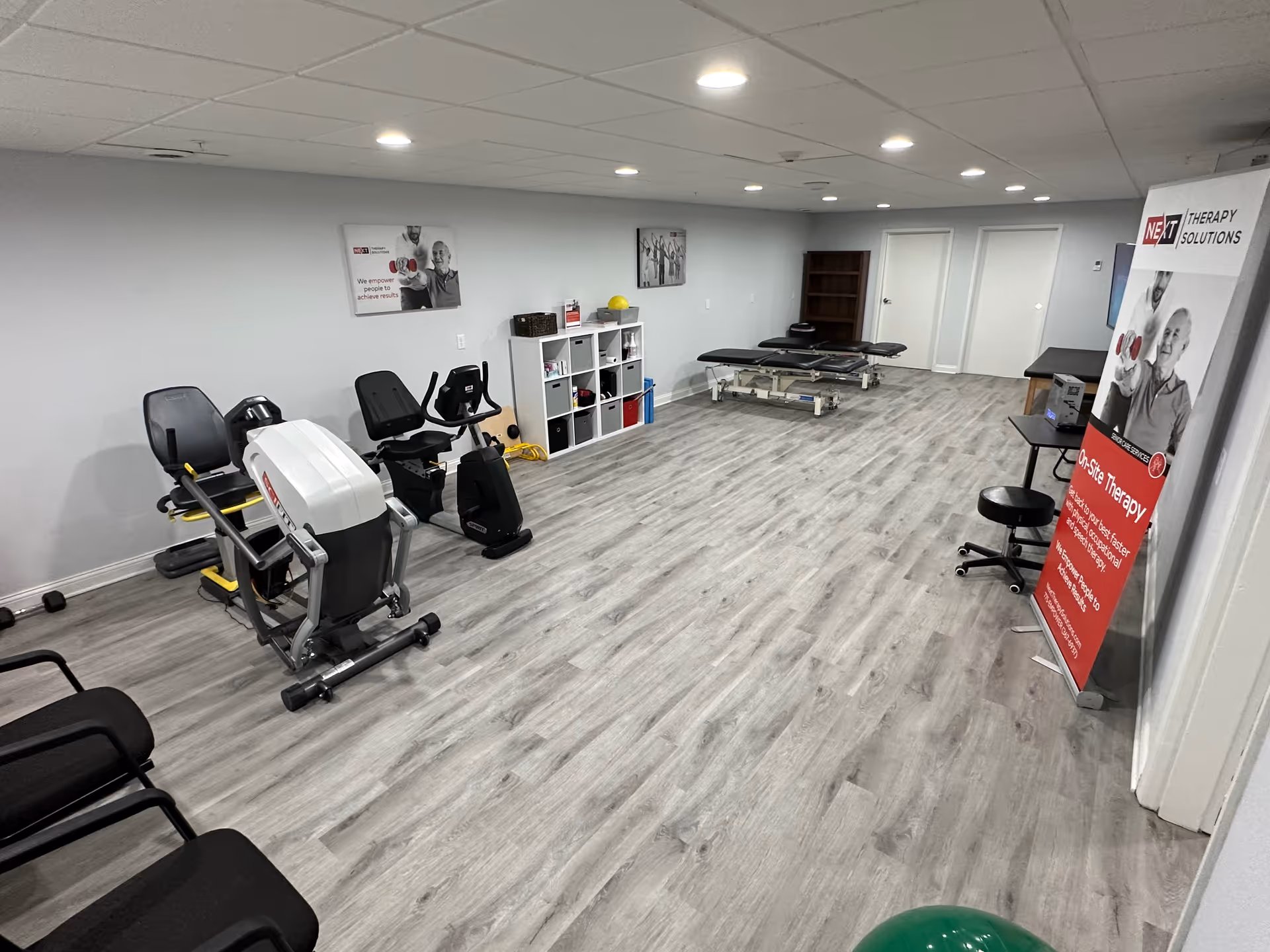 Therapy and exercise room with recumbent exercise machines, treatment tables, shelving, and a promotional banner.