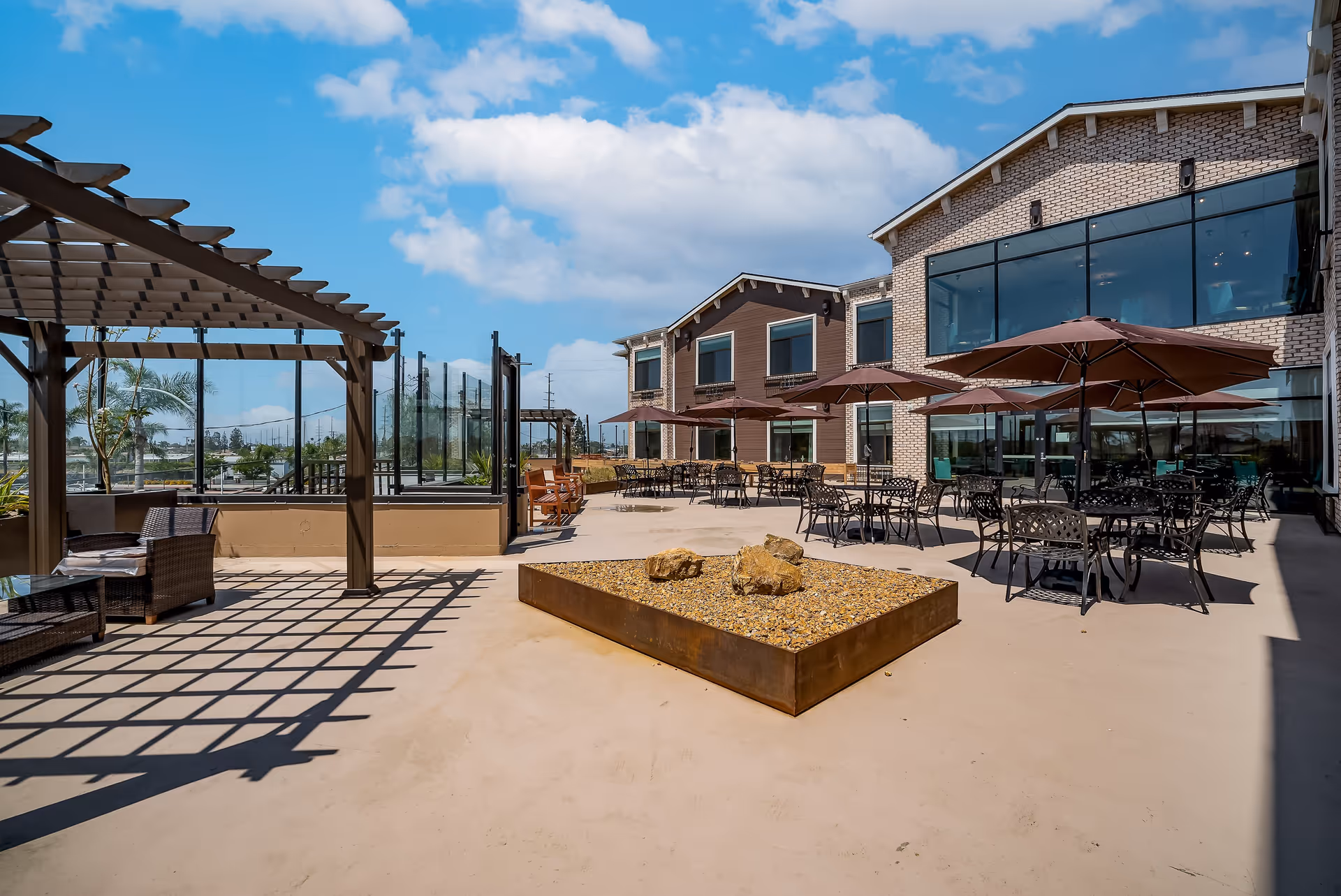 Outdoor patio area at Beach Terrace with several tables and chairs under large umbrellas, a pergola with seating, and a raised planter with rocks. The building has large windows and a brick and wood exterior under a partly cloudy blue sky.