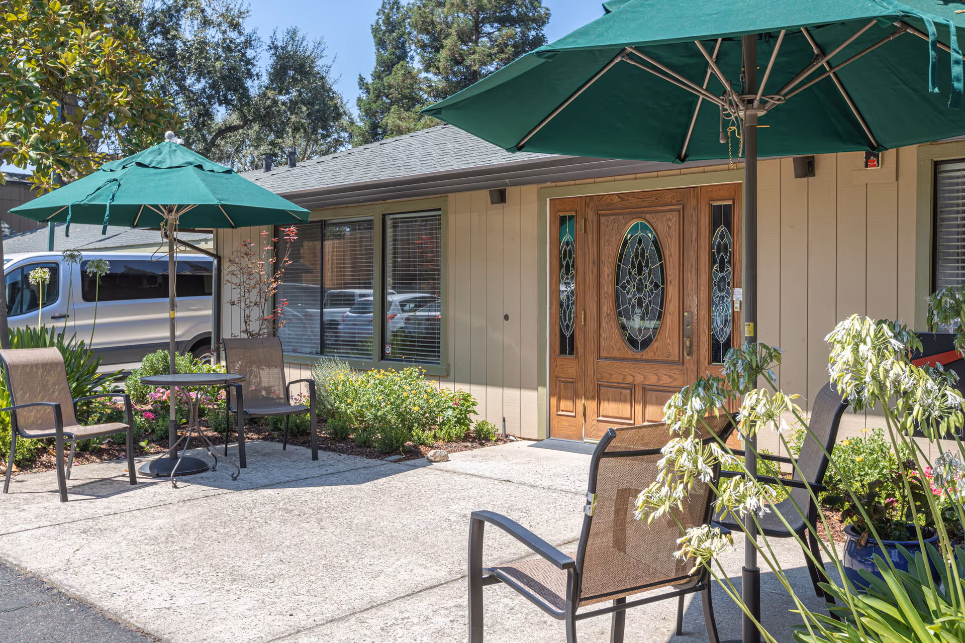 Outdoor seating area at Almond Grove Assisted Living featuring two green patio umbrellas, several brown mesh chairs, a small round table, and various plants and flowers near the building entrance with decorative wooden double doors and windows with blinds.