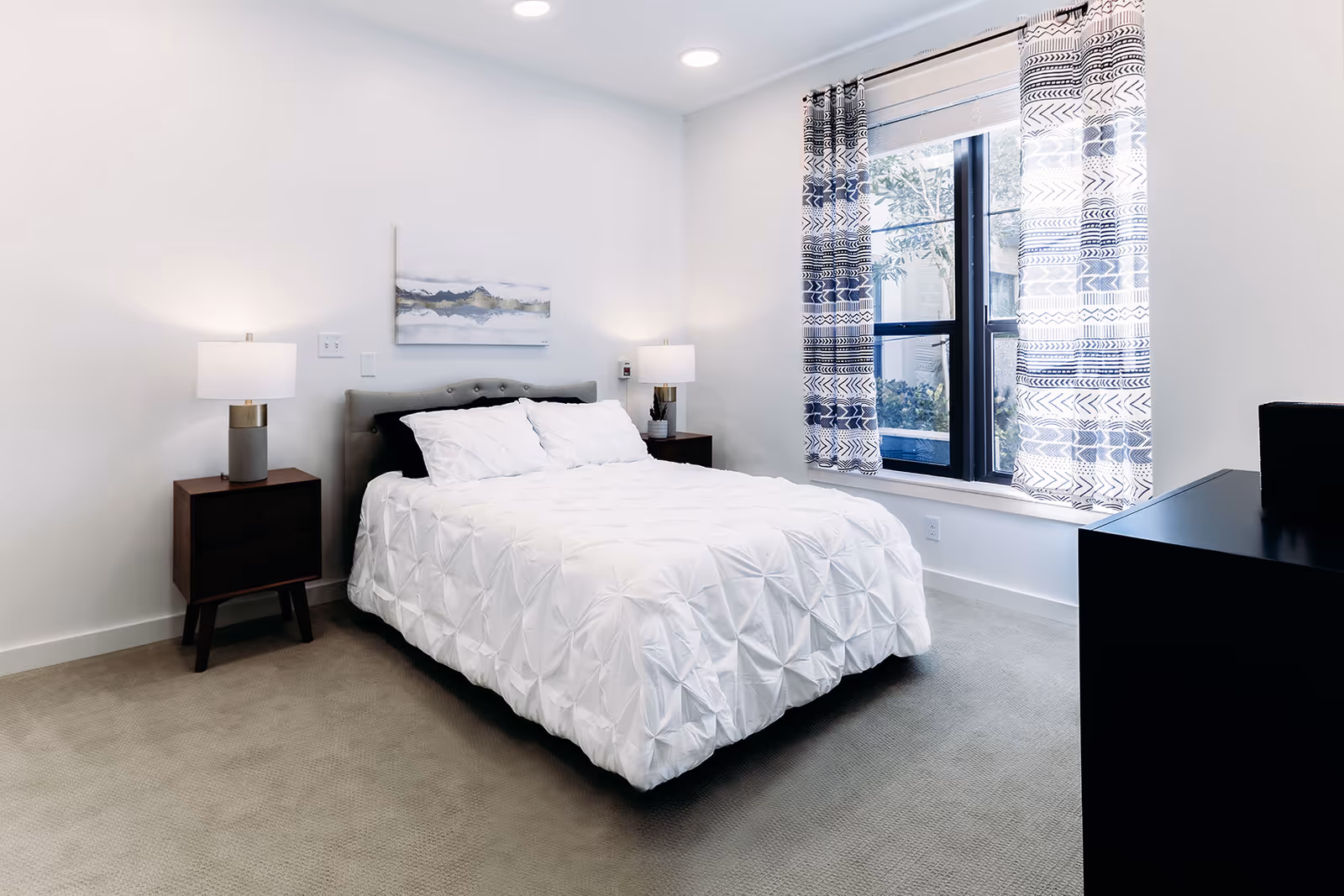 Bright modern bedroom with a neatly made white bed, two bedside tables with lamps, a window with patterned curtains, and a dresser.