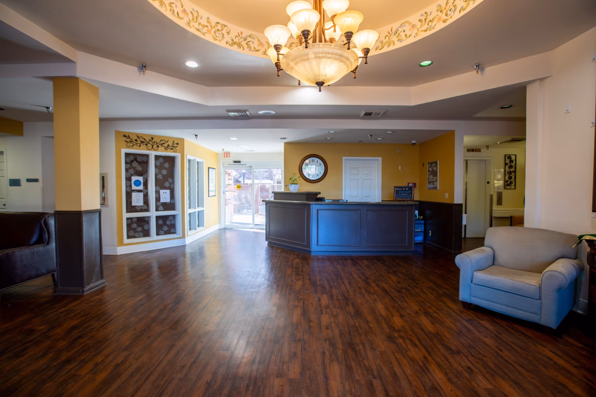 Spacious reception area of Lexington Assisted Living with wooden flooring, a dark wooden reception desk, a large round clock on the wall behind the desk, a comfortable armchair to the right, and a chandelier hanging from the ceiling. The walls are painted yellow and white with decorative accents, and there is a glass door entrance in the background.