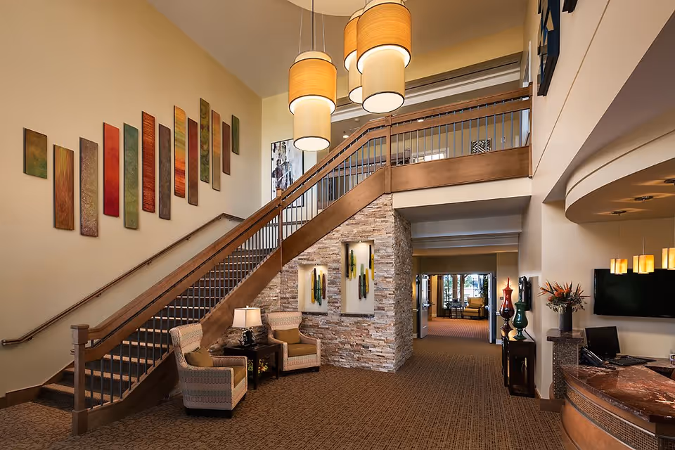 Interior view of a senior living facility lobby with a staircase featuring wooden handrails and metal balusters. Two armchairs with a small table and lamp are placed under the staircase. The walls are decorated with colorful vertical art panels and stone accents. Pendant lights hang from the ceiling, and a reception desk with a computer and decorative vases is visible on the right side. A hallway leads to another seating area in the background.