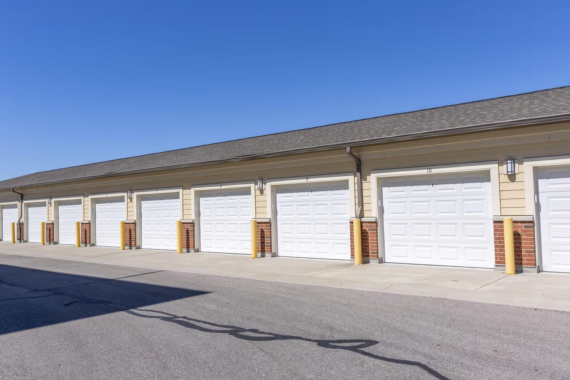 A row of closed white garage doors attached to a beige building with a shingled roof under a clear blue sky.