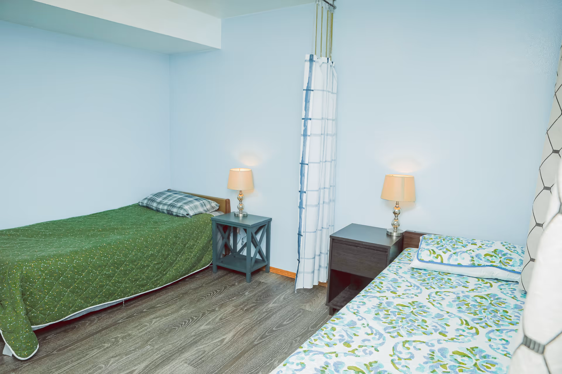 A small bedroom with two single beds placed against opposite walls. One bed has a green quilt and a plaid pillow, while the other has a floral patterned bedspread and matching pillow. Each bed has a nightstand with a lamp. The room has light blue walls, wood flooring, and a white curtain hanging in the corner.