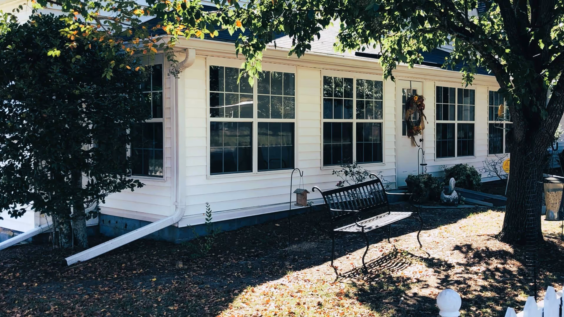 Exterior view of a white building with multiple large windows and a door decorated with a wreath. There is a black metal bench on the ground near the building, surrounded by trees and some garden decorations. Sunlight casts shadows on the ground covered with fallen leaves.