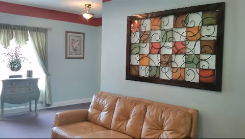 A cozy living room area with a tan leather couch against a light blue wall. Above the couch is a large decorative wall art piece featuring a grid of colorful squares with black wrought iron scrollwork. To the left, a window with sheer green curtains lets in natural light, and beneath the window is a small blue chest of drawers with decorative items on top. A framed floral picture hangs on the wall near the window.