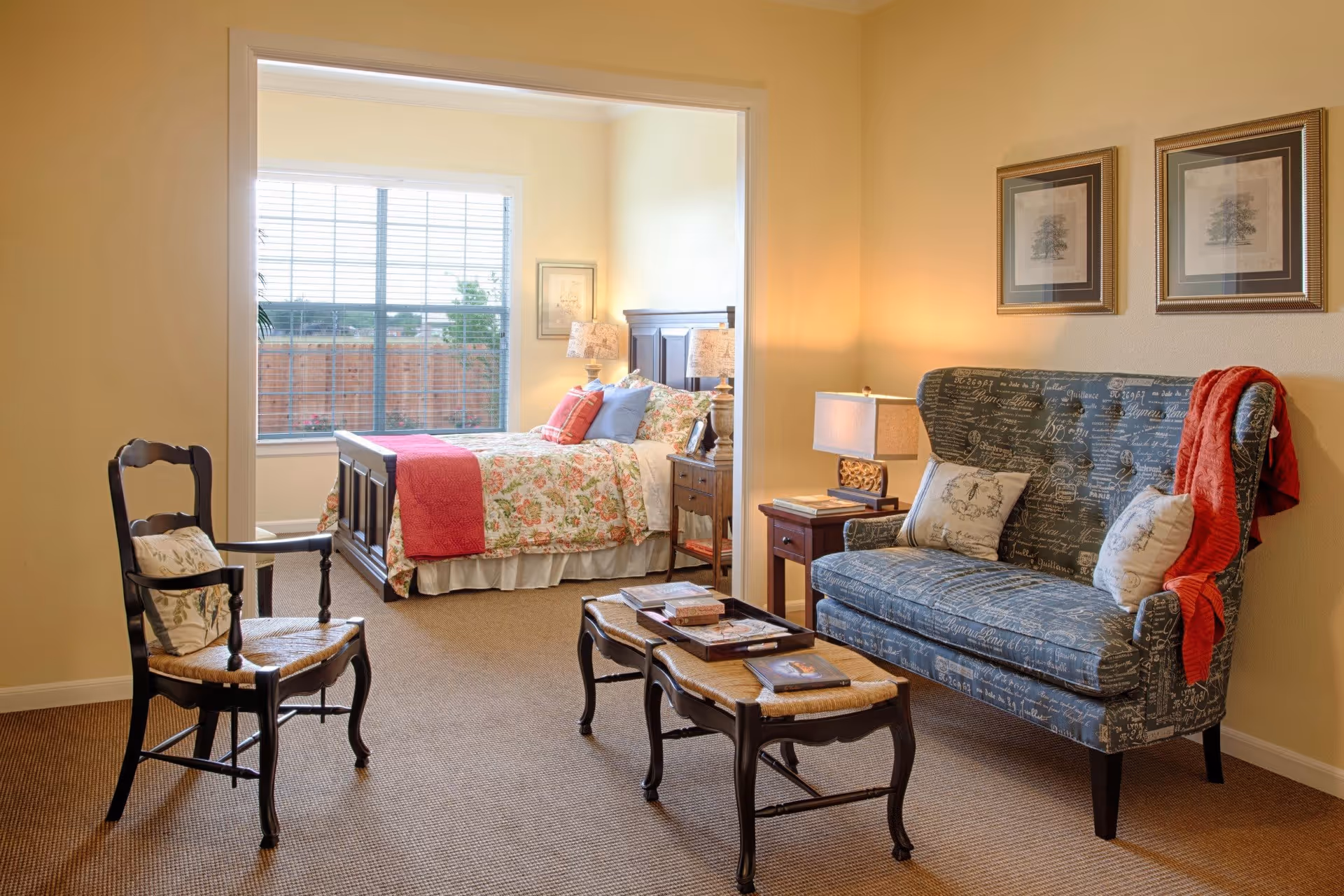 A cozy living area with a patterned blue loveseat adorned with two pillows and a red throw blanket. A wooden coffee table with books and magazines sits in front of the loveseat. To the left, there is a wooden chair with a cushion. In the background, an adjoining bedroom is visible with a bed covered in floral bedding and a red throw blanket, a nightstand with a lamp, and a large window with blinds letting in natural light.