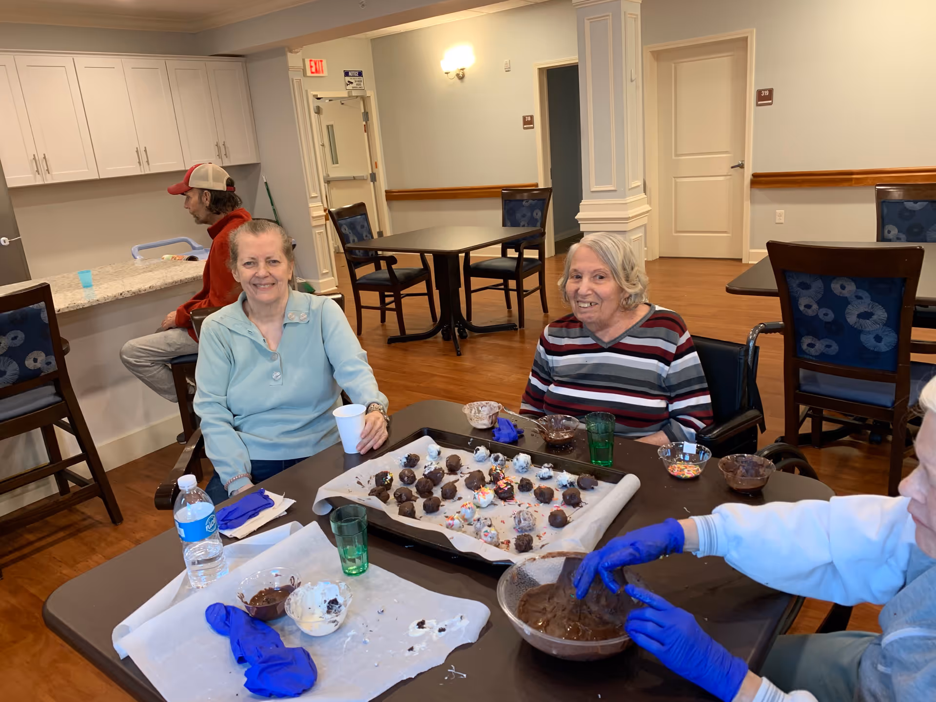 Three elderly individuals sitting around a table in a common area of a senior living facility. Two women are smiling at the camera while sitting at the table with trays of chocolate-covered treats and bowls of ingredients. One person is wearing blue gloves and appears to be dipping treats in chocolate. In the background, a man in a red jacket and cap is seated at a counter. The room has wooden floors, light-colored walls, and several tables and chairs.