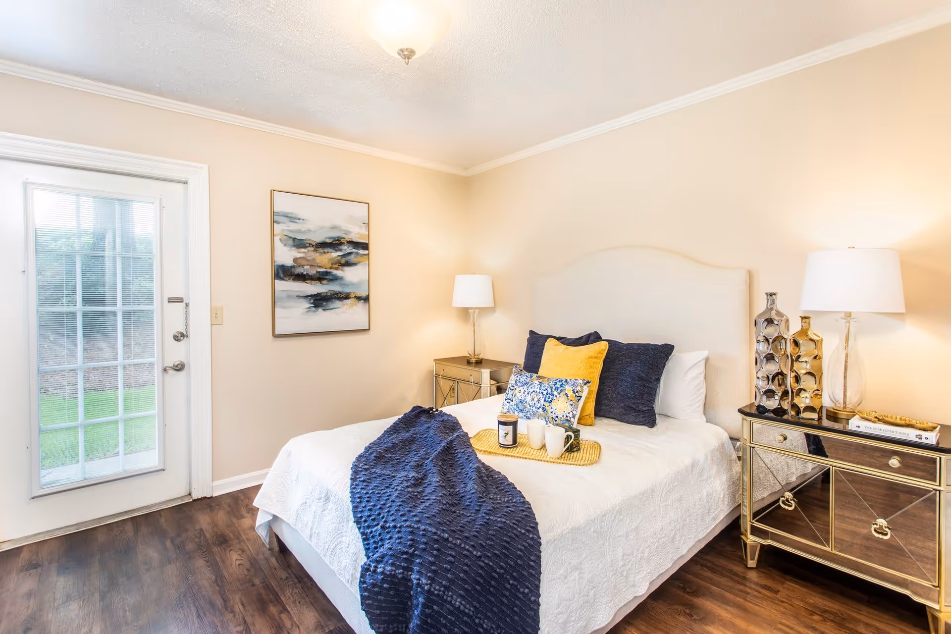A bright bedroom with a white bed covered with a white quilt and decorated with navy blue and yellow pillows. A navy blue textured throw blanket is draped over the bed. There are two mirrored nightstands on either side of the bed, each with a lamp and decorative items. A framed abstract painting hangs on the wall, and a glass door leads outside to a green lawn.