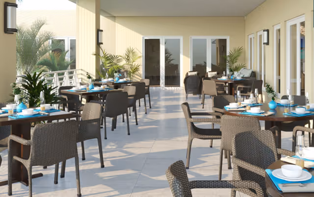 Outdoor covered patio dining area with multiple tables set with plates, bowls, glasses, and blue placemats. Wicker chairs surround the tables, and potted plants are placed along the walls and railing. The area is well-lit with natural light and has beige walls and tiled flooring.