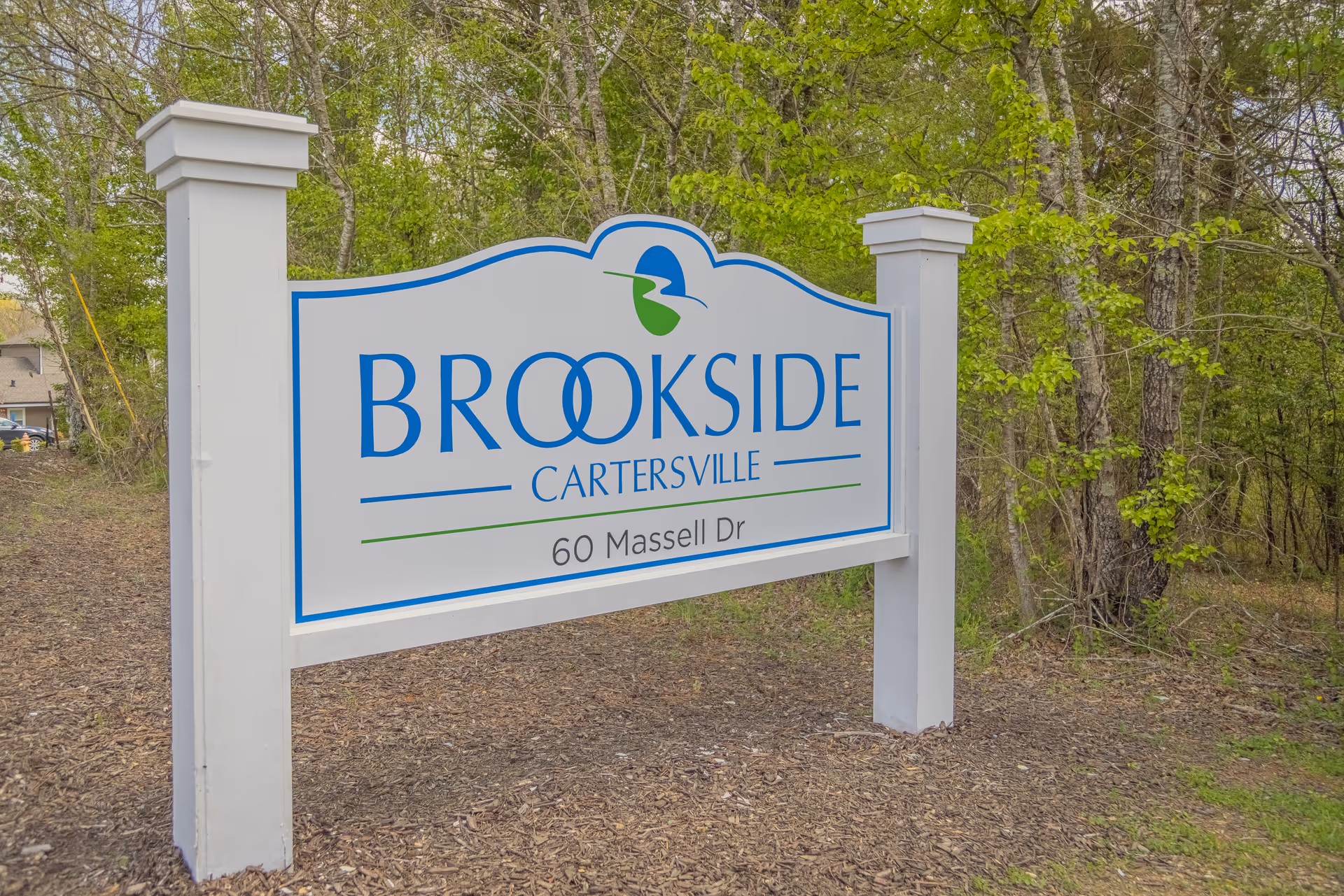 A white and blue sign for Brookside Cartersville senior living facility located at 60 Massell Dr, set outdoors with trees and greenery in the background.