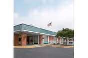 Exterior view of Charlottesville Health & Rehabilitation Center showing a single-story building with a green metal roof, brick pillars, an American flag on a flagpole, and a paved driveway in front.