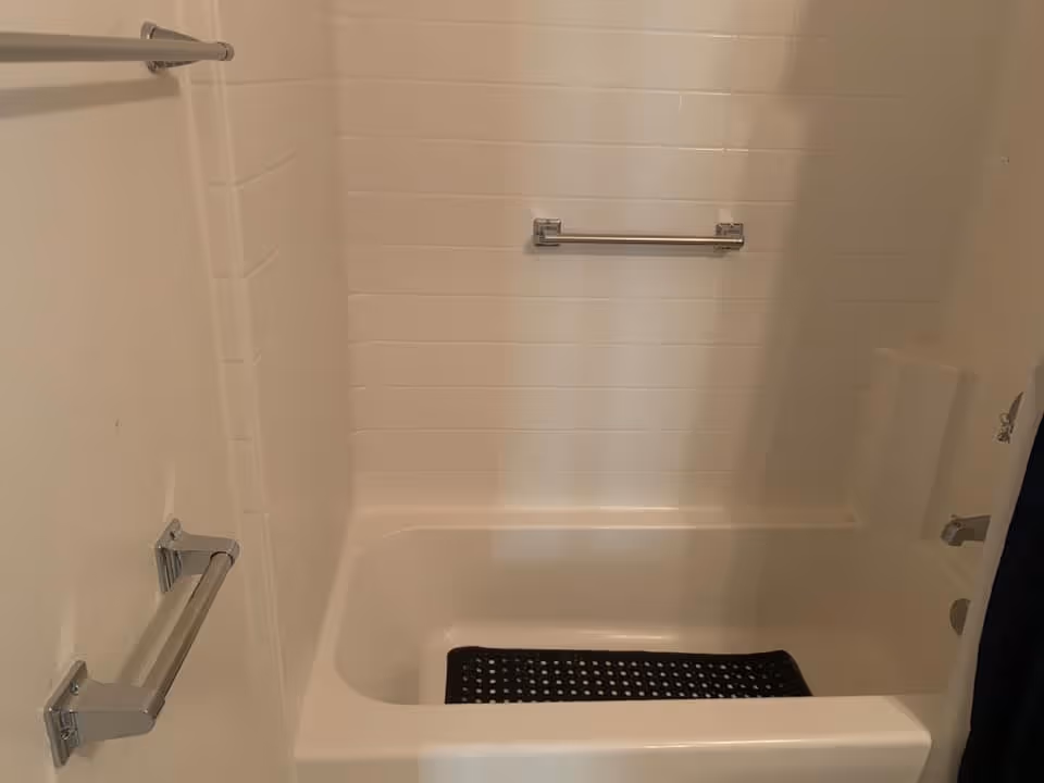 A white bathtub with a black non-slip mat inside, surrounded by white tiled walls. There are two silver grab bars on the left wall and one silver grab bar on the back wall above the tub.
