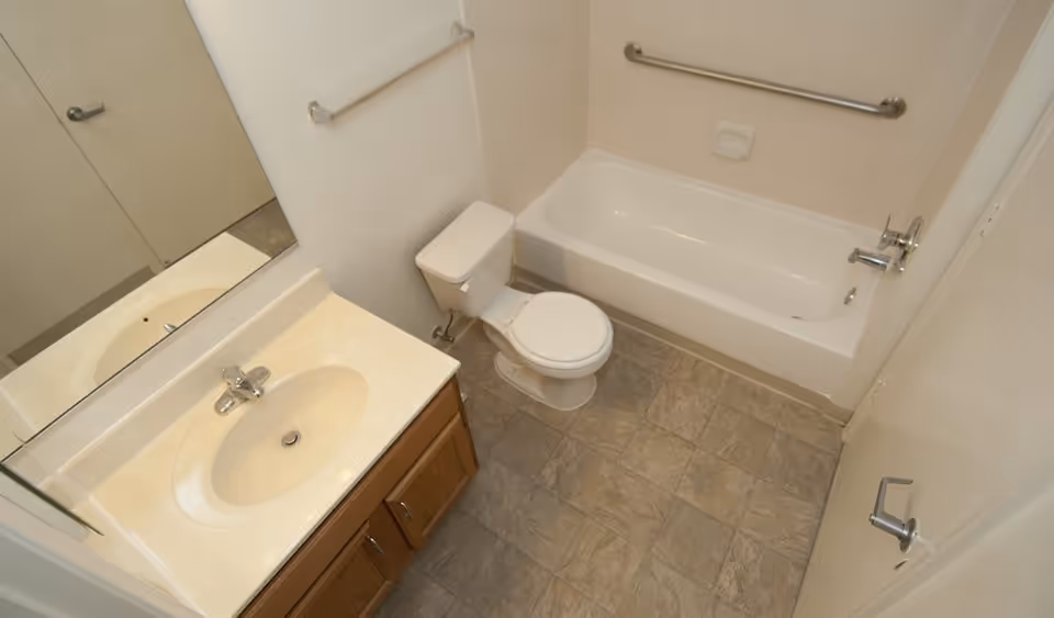 A clean bathroom with a white bathtub, a toilet, a sink with a countertop and wooden cabinet, a large mirror above the sink, and a tiled floor. There are grab bars installed on the wall near the bathtub and above the toilet.