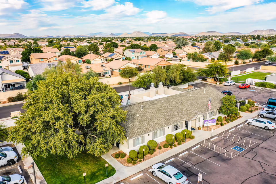 Aerial view of a single-story building with a gray roof surrounded by neatly trimmed bushes and a large tree in front. The building is adjacent to a parking lot with several parked cars and a visible handicap parking space. In the background, there is a residential neighborhood with many houses and distant mountains under a partly cloudy sky.