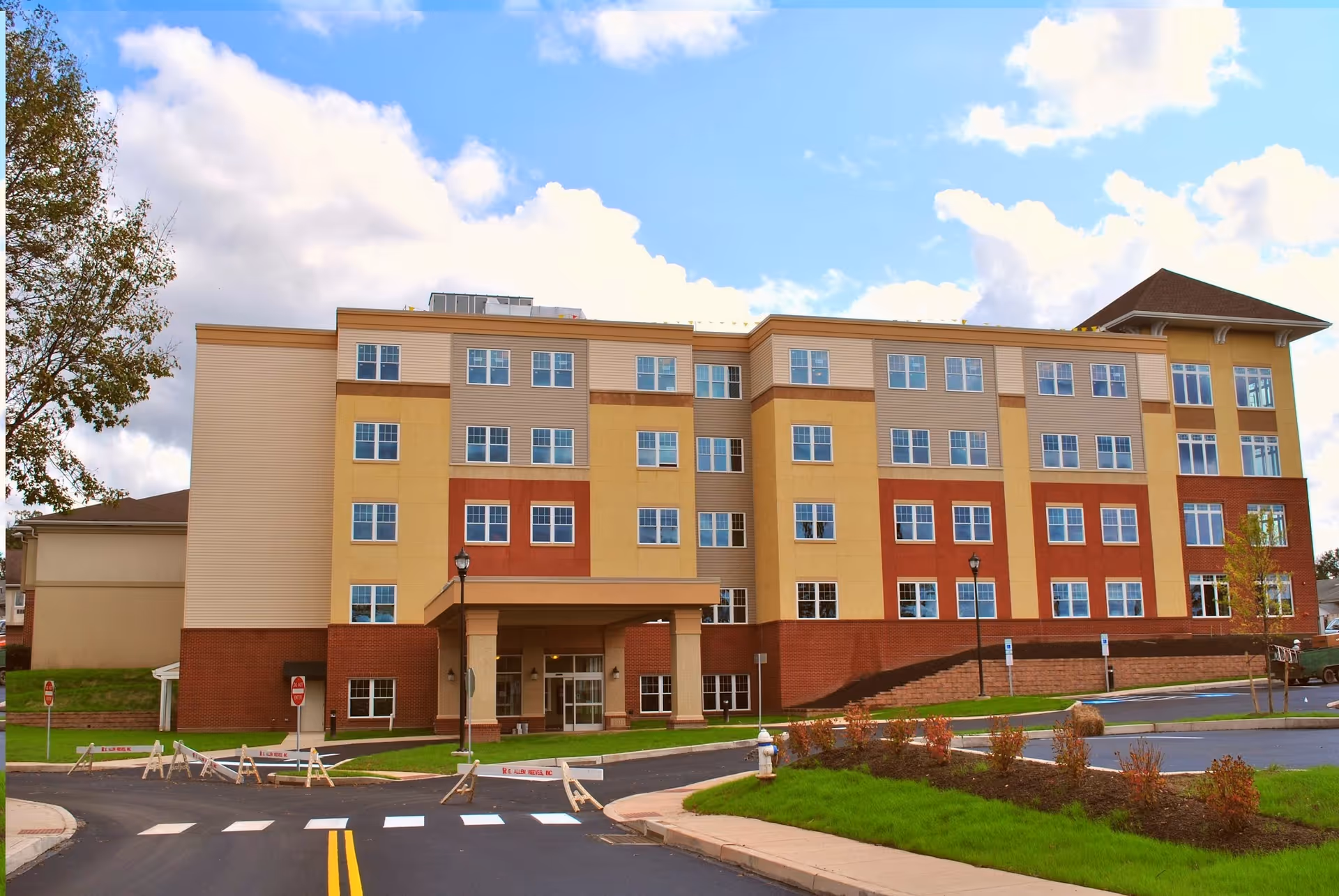 Exterior view of a multi-story senior living facility building with a covered entrance, multiple windows, and a landscaped area with grass and small shrubs in front. The sky is partly cloudy.